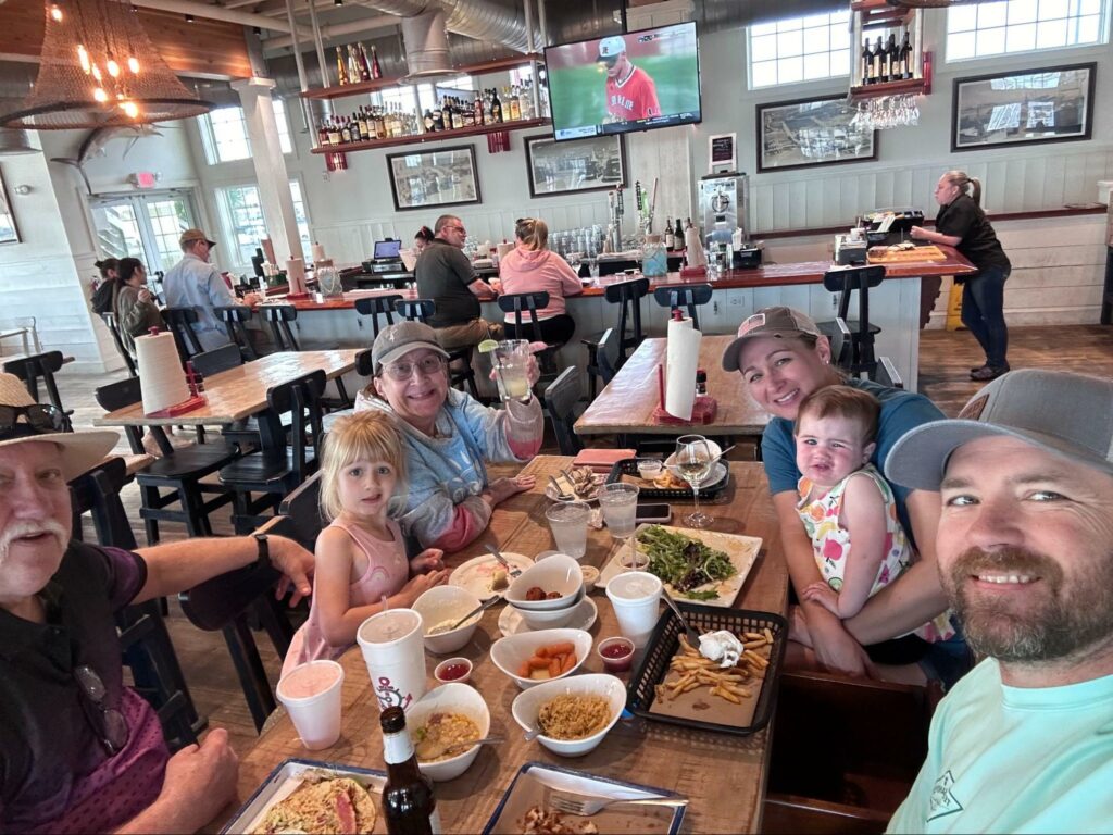 A family enjoying a meal together inside Grumble’s Seafood Co., featuring six members including two children and four adults, seated at a large table filled with various seafood dishes. The restaurant has a coastal decor, with a bar area in the background where a baseball game is displayed on a television.