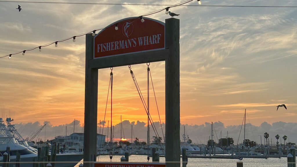 A picturesque sunset view at Fisherman's Wharf, featuring a large sign with the wharf's name on a wooden post, string lights, birds in flight, and a backdrop of a vibrant sky over a marina filled with boats.