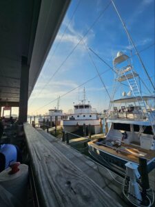 View from the patio of Grumble’s Seafood Co., featuring a glimpse of the marina with several boats moored at the docks, including a large yacht and a fishing boat. The perspective is framed by the restaurant’s covered deck, showcasing the calm ocean and clear sky in the background.